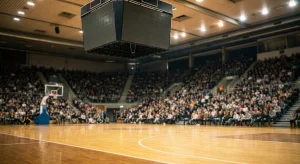 Empty hardwood basketball court with a blank jumbotron hanging above. Blurred crowds fill the surrounding stadium seats